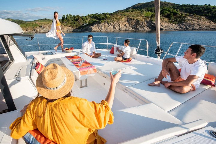 People relaxing on a yacht deck near a rocky coastline.