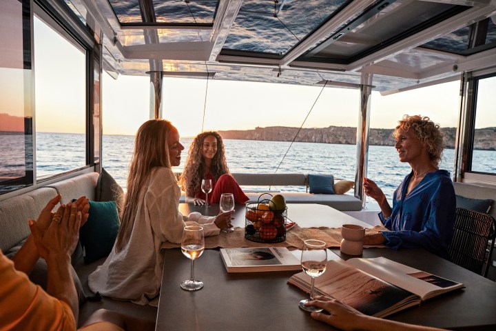 Four people sitting at a table in a boat, laughing and enjoying drinks at sunset.