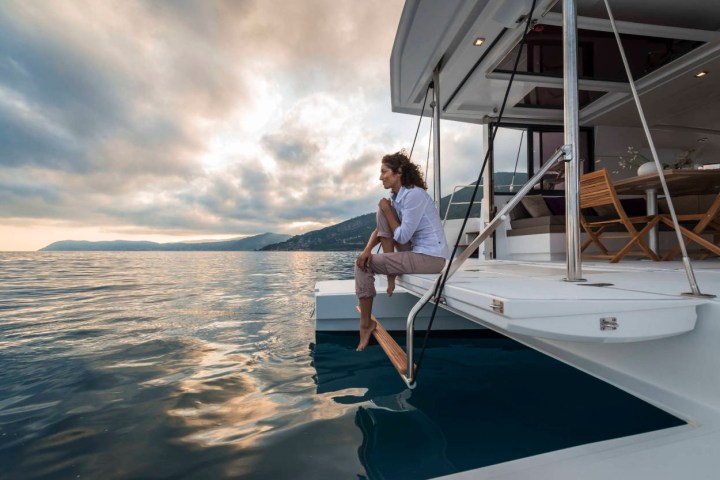 Person sitting on yacht deck, feet touching water, with cloudy sky and mountains in the background.